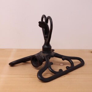 May include: A black metal lawn sprinkler featuring a tripod base with decorative scrollwork and a central rotating head. The device includes a threaded circular intake port at the front for hose attachment, constructed with a sturdy, dark finish.