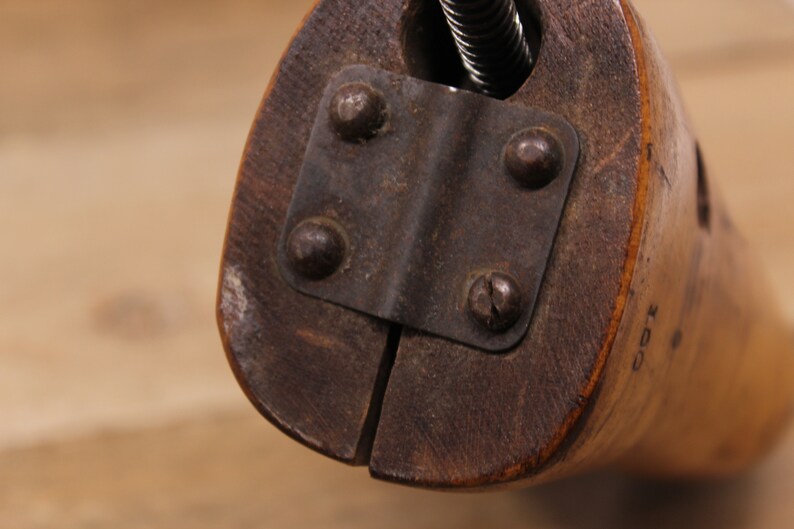 May include: Close-up of a wooden shoe stretcher with a metal clamp and screws. The stretcher has a brown finish and is made of wood. The clamp is made of metal and has a black finish.