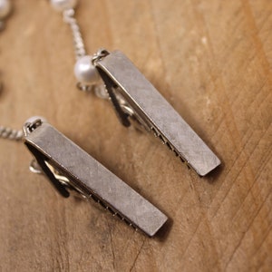 May include: Two silver metal hair clips with a chain and white pearls attached. The clips are lying on a wooden surface.