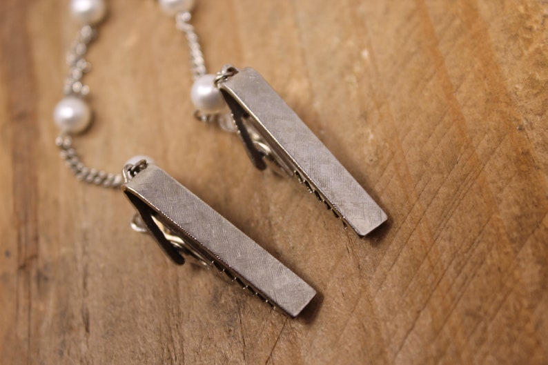 May include: Two silver metal hair clips with a chain and white pearls attached. The clips are lying on a wooden surface.