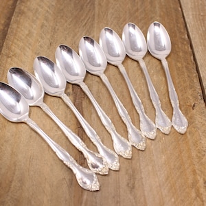 May include: Eight silver spoons arranged diagonally on a wooden surface. The spoons have a classic design with ornate detailing on the handles and a polished, reflective finish. The spoons are likely for serving or eating.