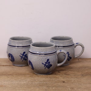 May include: Three blue and white ceramic mugs with a speckled glaze and blue floral designs. The mugs have a rounded body and a handle.