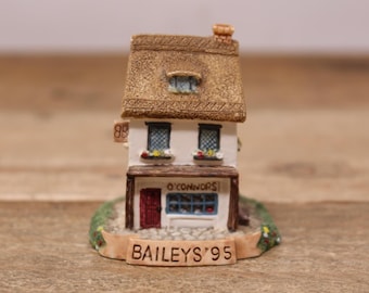 May include: A miniature ceramic replica of a pub with a thatched roof. The building is white with a red door and blue windows. The sign reads "O'Connors" and "Baileys '95". The base is green with small flowers.