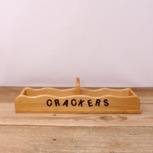 May include: A light-colored wooden cracker serving tray with a handle. The word "CRACKERS" is printed in black on the front. The tray has a wavy top edge and is sitting on a wooden surface.