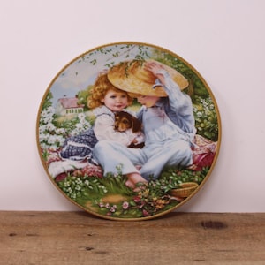 May include: A porcelain plate featuring a painted image of two children sitting in a field with a puppy. The girl is wearing a blue and white dress and the boy is wearing overalls. They are both holding the puppy. The plate has a gold rim.