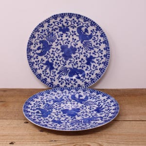 May include: Two round ceramic plates featuring a white base with a detailed blue pattern of stylised birds and floral motifs. The design includes a decorative border and a central arrangement of swirling vines, leaves, and bird figures in blue.
