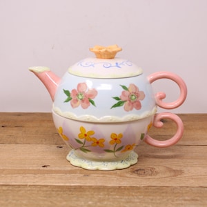 May include: A pastel pink and blue teapot with a floral design. The teapot has a matching cup and saucer. The teapot has a lid with a flower on top.