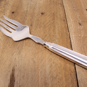 May include: A silver serving fork with four prongs and a long, rectangular handle. The fork is made of shiny metal and rests on a wooden surface. The handle has a decorative, ridged design near the fork's head.
