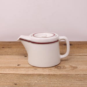 May include: A white ceramic teapot with a lid, featuring a red and green stripe design around the rim and lid. The teapot has a curved handle and a spout for pouring. It sits on a wooden surface.