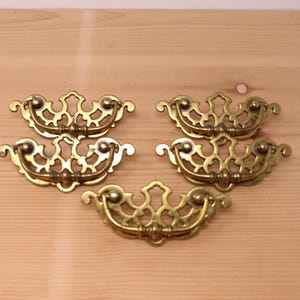 May include: Five brass drawer pulls featuring an ornate, openwork backplate with a symmetrical, scrolling design and a matching swinging bail handle. Each piece has a polished metallic finish with intricate cutouts and curved edges.
