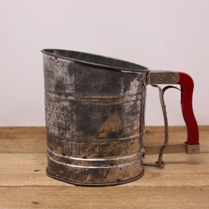 May include: Vintage metal flour sifter with a red handle. The sifter has a worn, aged look and is made of metal with a mesh screen.
