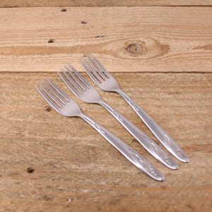May include: Three stainless steel forks with a floral pattern on the handles, set diagonally on a wooden surface. Each fork has four tines and a polished finish, ideal for a dining setting.