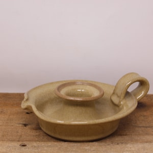 May include: A light brown ceramic candle holder with a single candle well and a handle. The holder has a textured surface and a slightly rounded shape.