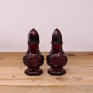 May include: Two antique ruby red glass salt and pepper shakers. Each shaker has a faceted, pointed top with small holes and a decorative, embossed pattern on the body. The shakers are on a wooden surface.