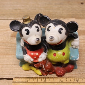 May include: Vintage ceramic figurine of Mickey Mouse and Minnie Mouse sitting on a blue bench. Mickey is wearing a red and white polka dot shirt and Minnie is wearing a yellow and green polka dot dress.