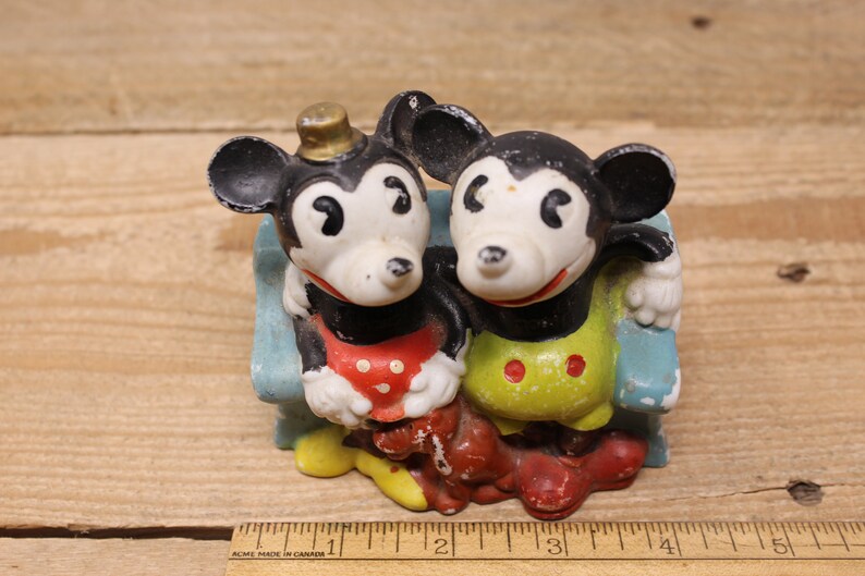 May include: Vintage ceramic figurine of Mickey Mouse and Minnie Mouse sitting on a blue bench. Mickey is wearing a red and white polka dot shirt and Minnie is wearing a yellow and green polka dot dress.