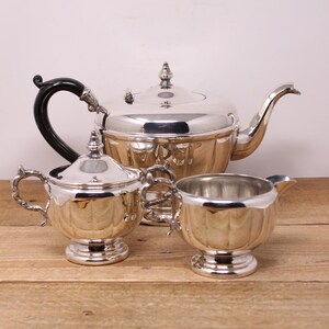May include: A silver tea set with a teapot, creamer, and sugar bowl. The teapot has a black handle and a shiny, reflective finish. The creamer and sugar bowl have ornate handles.