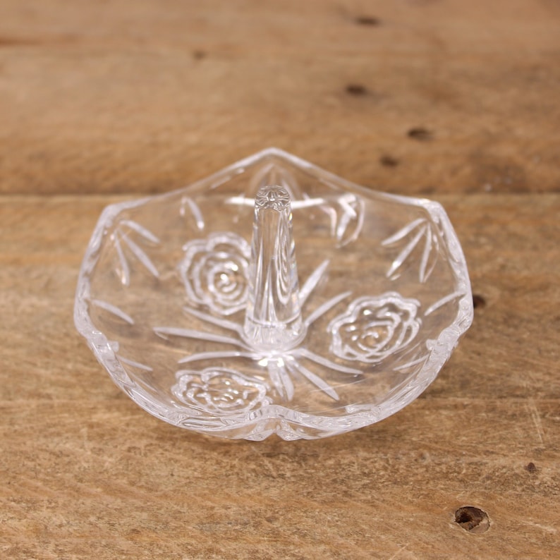 May include: A clear glass ring holder with a raised center post and a floral design. The holder is shaped like a flower with a scalloped edge.