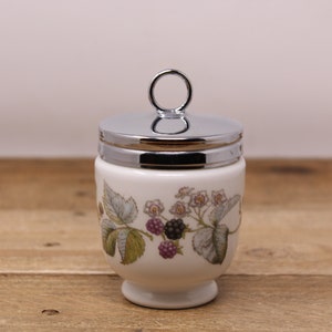 May include: White ceramic egg cup with a silver lid and a floral design featuring blackberries and leaves. The lid has a ring handle.