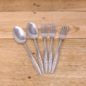 May include: A collection of stainless steel cutlery, comprising two spoons and three forks, displayed on a wooden surface. The cutlery has a floral pattern on the handles.