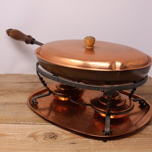 May include: Copper chafing dish with a hammered finish, oval pan, and wooden handle. The dish sits on a wrought iron stand above two burners, all on a matching copper tray. The lid has a wooden knob.
