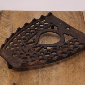 May include: A vintage cast iron trivet with a decorative design. The trivet is shaped like a traditional iron and has a circular opening in the centre.