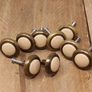 May include: A collection of ten antique-style cabinet knobs. Each knob features a cream-colored center, a brass-toned rim, and a silver-colored screw. The knobs are arranged on a weathered wooden surface.