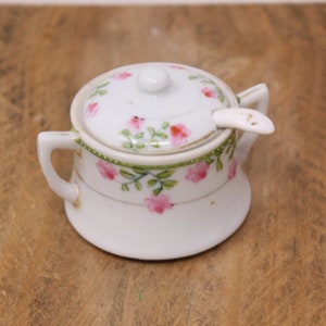May include: A white porcelain sugar bowl with a lid and a small spoon. The bowl and lid are decorated with pink flowers and green leaves.