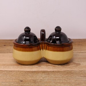 May include: A two-section ceramic condiment server with a handle. The server has a light yellow base, a brown middle section, and a dark brown lid. The lids have a small knob on top. The server is on a wooden surface.