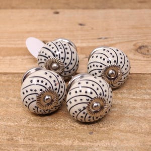 May include: Four white ceramic cabinet knobs with a black spiral and dot pattern. Each knob has a silver-coloured metal base and a decorative silver-coloured flower-shaped accent. The knobs are on a light brown wooden surface.