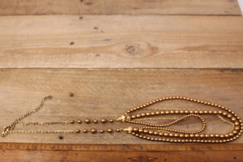 May include: A gold multi-strand necklace with round beads. The necklace is made of multiple strands of gold chain and gold beads.