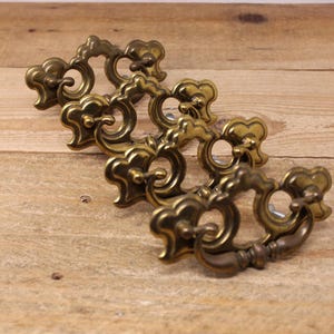 May include: Four antique brass drawer pulls with ornate, floral-inspired designs. The pulls have a warm, aged patina and a ring-shaped handle. They are arranged on a light brown wooden surface, showcasing their intricate details and vintage aesthetic.