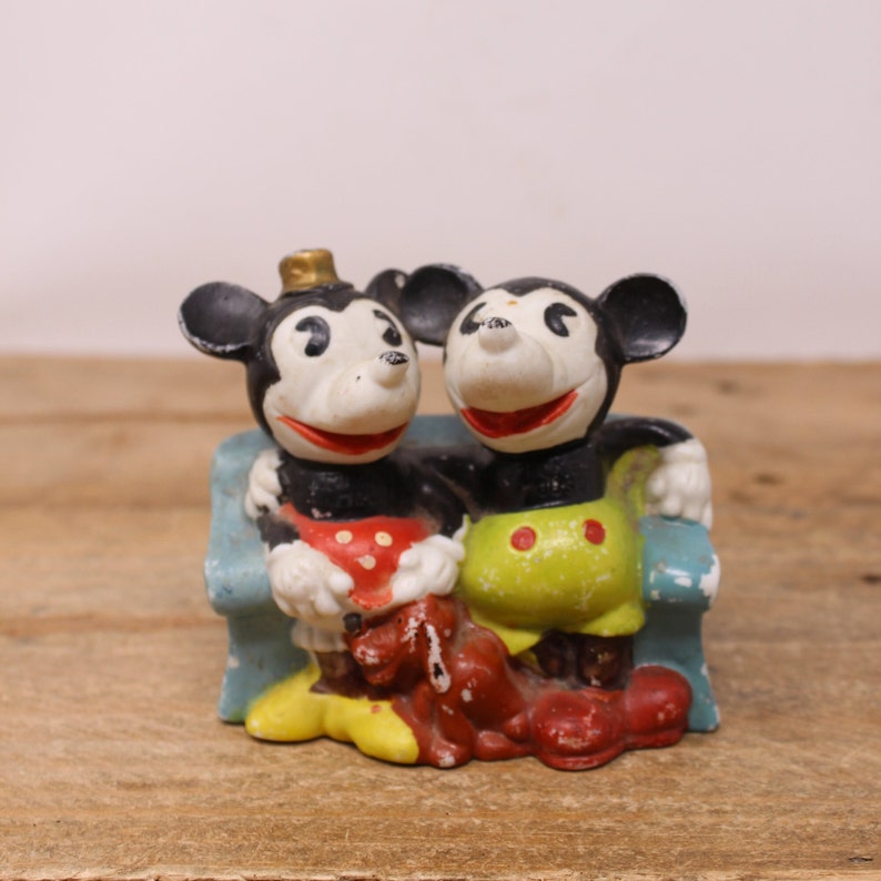 May include: A vintage ceramic figurine of Mickey Mouse and Minnie Mouse sitting on a blue bench. Mickey is wearing a red and white polka dot shirt and Minnie is wearing a yellow and white polka dot shirt.