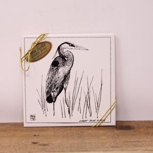 May include: Square ceramic tile with a black and white illustration of a Great Blue Heron in tall grass. The tile is bordered and labelled "Nova Scotia Wildlife Series". A gold-coloured wire holds a tag.