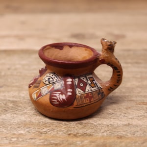 May include: A small, hand-painted ceramic vessel with a handle and spout. The vessel is primarily tan with a dark brown rim and handle. It features geometric patterns in red, white, and black, showcasing a traditional design.