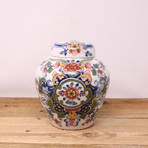 May include: A white ceramic jar with a lid, adorned with a vibrant floral pattern in red, blue, yellow, and green. The jar has a rounded shape and sits on a wooden surface. The lid mirrors the floral design.