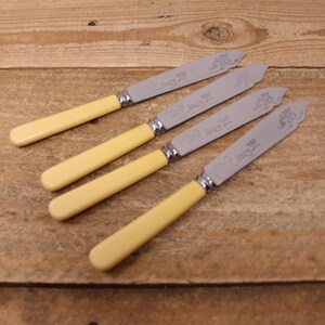 May include: Four butter knives with cream-colored handles and silver blades. The blades have a decorative floral design. The knives are arranged diagonally on a wooden surface.