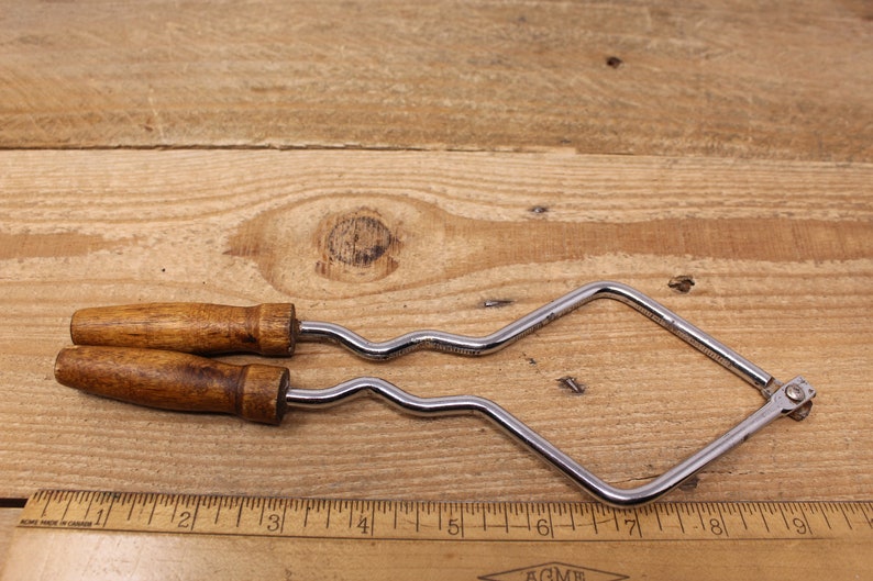 May include: A pair of vintage metal and wood jig saw frames with a wavy blade. The frames are silver and the handles are brown wood. The frames are about 7 inches long.