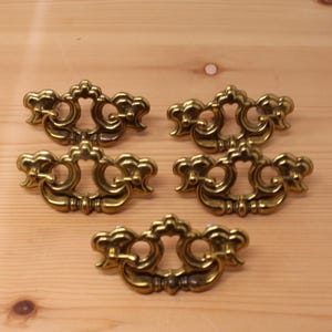 May include: Five brass-toned metal drawer pulls arranged in a group. Each pull features an ornate, symmetrical design with a central openwork plate and a swinging bail handle attached to two side posts, showcasing a polished metallic finish.