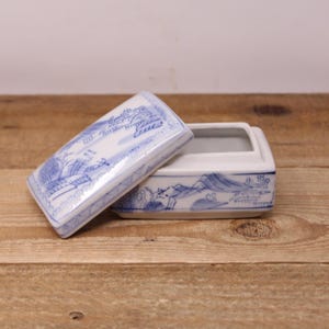 May include: A rectangular white ceramic trinket box with a lid, decorated with blue landscape illustrations. The lid is slightly open, revealing the interior. The box and lid feature detailed blue drawings.