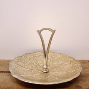 May include: A decorative, leaf-shaped serving tray with a silver handle. The tray has a textured, off-white surface and a silver rim. The handle is a simple, arched design. The tray is likely made of metal.