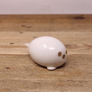 May include: A small, white ceramic seal figurine with three brown spots and a small tail. The seal is oval-shaped and sits on a wooden surface. The figurine is a decorative item.