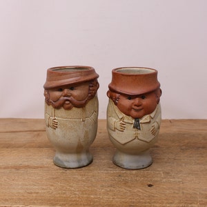 May include: Two ceramic mugs shaped like men with brown hats and facial features. The mugs are glazed in a light brown and white color.