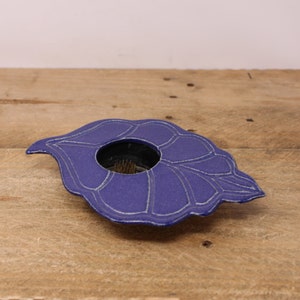 May include: A blue ceramic flower frog in a leaf shape with a central opening. The leaf design has white etched lines. The flower frog is designed to hold flowers in place for floral arrangements. The background is a wooden surface.