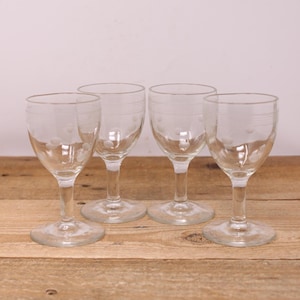 May include: Four clear glass cordial glasses with etched dot and line designs. The glasses have a rounded bowl, a slender stem, and a circular base. They are arranged on a wooden surface.