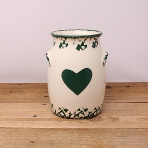 May include: Cream ceramic vase with a dark green heart and decorative green trim. The vase has two small handles and a slightly flared rim. The design is rustic and charming, suitable for farmhouse or country decor.