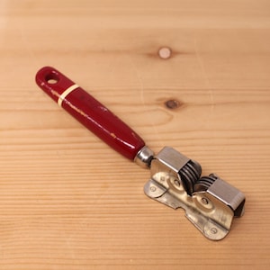 May include: A manual knife sharpener featuring a deep red handle with a cream-coloured band near the top and a hole for hanging. The sharpening mechanism consists of two sets of small, circular metal grinding wheels housed in a silver-toned frame.