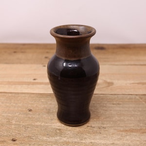 May include: A handcrafted ceramic vase with a dark brown rim and a glossy black body. The vase has a slightly flared opening and a rounded base. The exterior has a textured, ribbed design. The vase is displayed on a wooden surface.