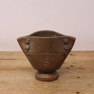 May include: A brown wooden bowl with a pedestal base. The bowl has carved geometric patterns and decorative elements. The design is reminiscent of rustic or tribal art. The bowl is likely handmade.