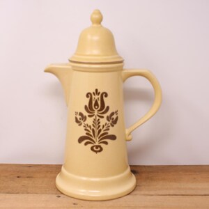 Peut inclure: Une cafetière en céramique jaune avec un motif floral brun. La cafetière a un couvercle et une poignée.